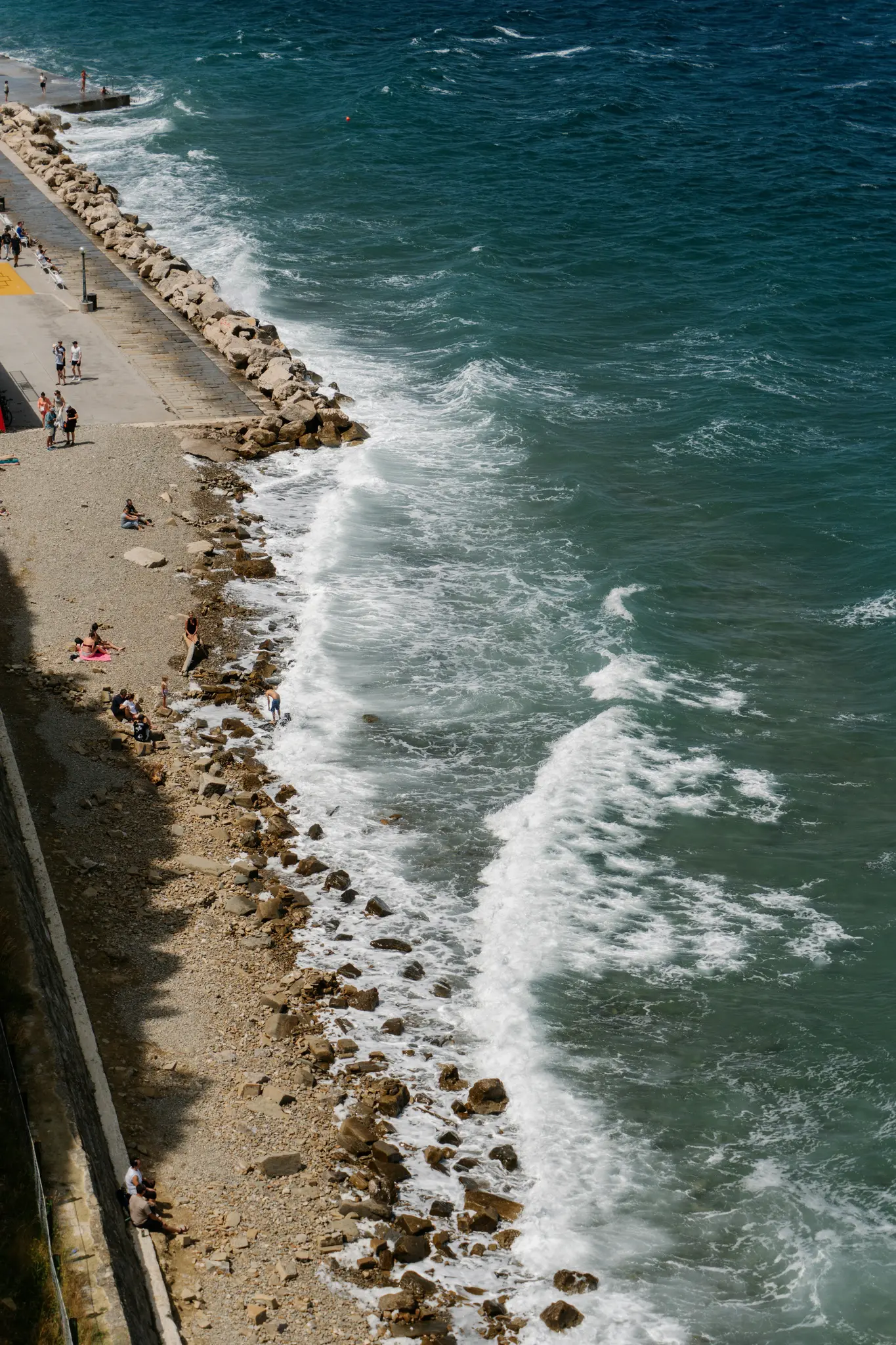 Een luchtfoto toont een levendige blauwgroene zee met witte golven die op een rotsachtig strand slaan. Een betonnen promenade