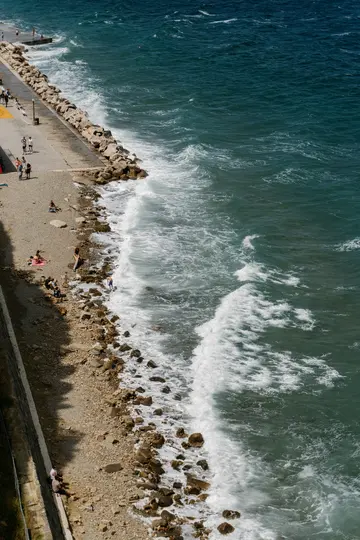 Een luchtfoto toont een levendige blauwgroene zee met witte golven die op een rotsachtig strand slaan. Een betonnen promenade