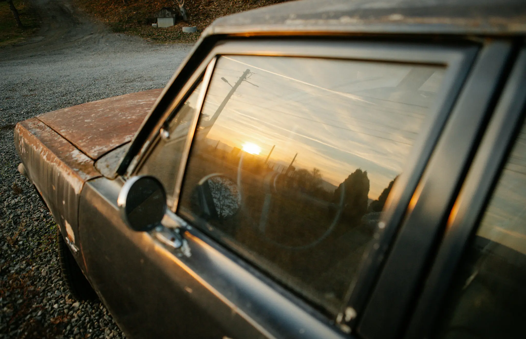 Een close-up van de zijkant van een oude, roestige auto. De ondergaande zon wordt fel gereflecteerd in het raam van de auto.