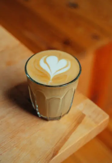 Un bicchiere di latte con un perfetto disegno a cuore bianco nella schiuma, posizionato su una superficie di legno chiaro.