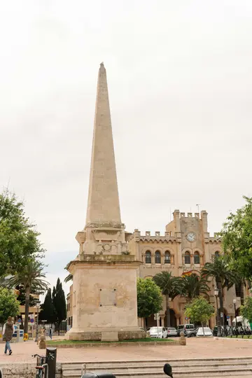 Vysoký, světlý kamenný obelisk stojí výrazně uprostřed náměstí za oblačného nebe. V pozadí jsou vidět budovy.