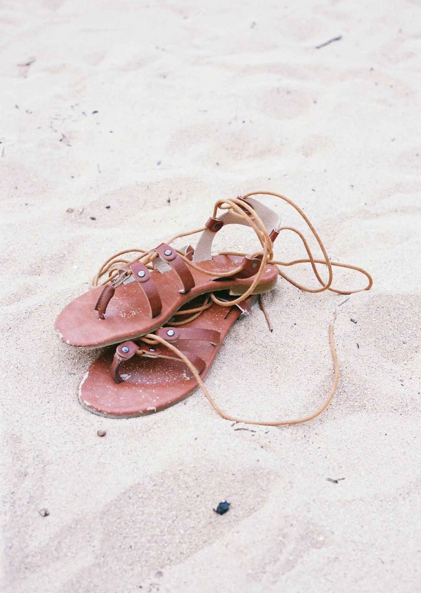 Een paar bruine leren sandalen met veters ligt op licht zand, met zandkorrels zichtbaar op het leer.