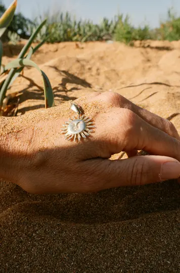 Una mano, parzialmente sepolta nella sabbia, tiene un ciondolo a forma di sole in argento con una faccia sorridente. Piante