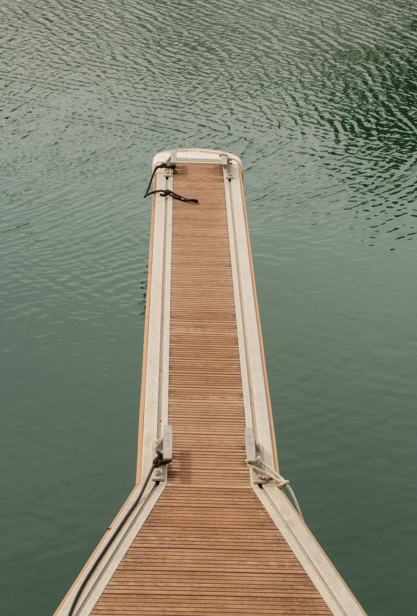 Vue en plongée d'une jetée étroite en bois aux bords gris clair s'avançant dans une eau calme et vert foncé.
