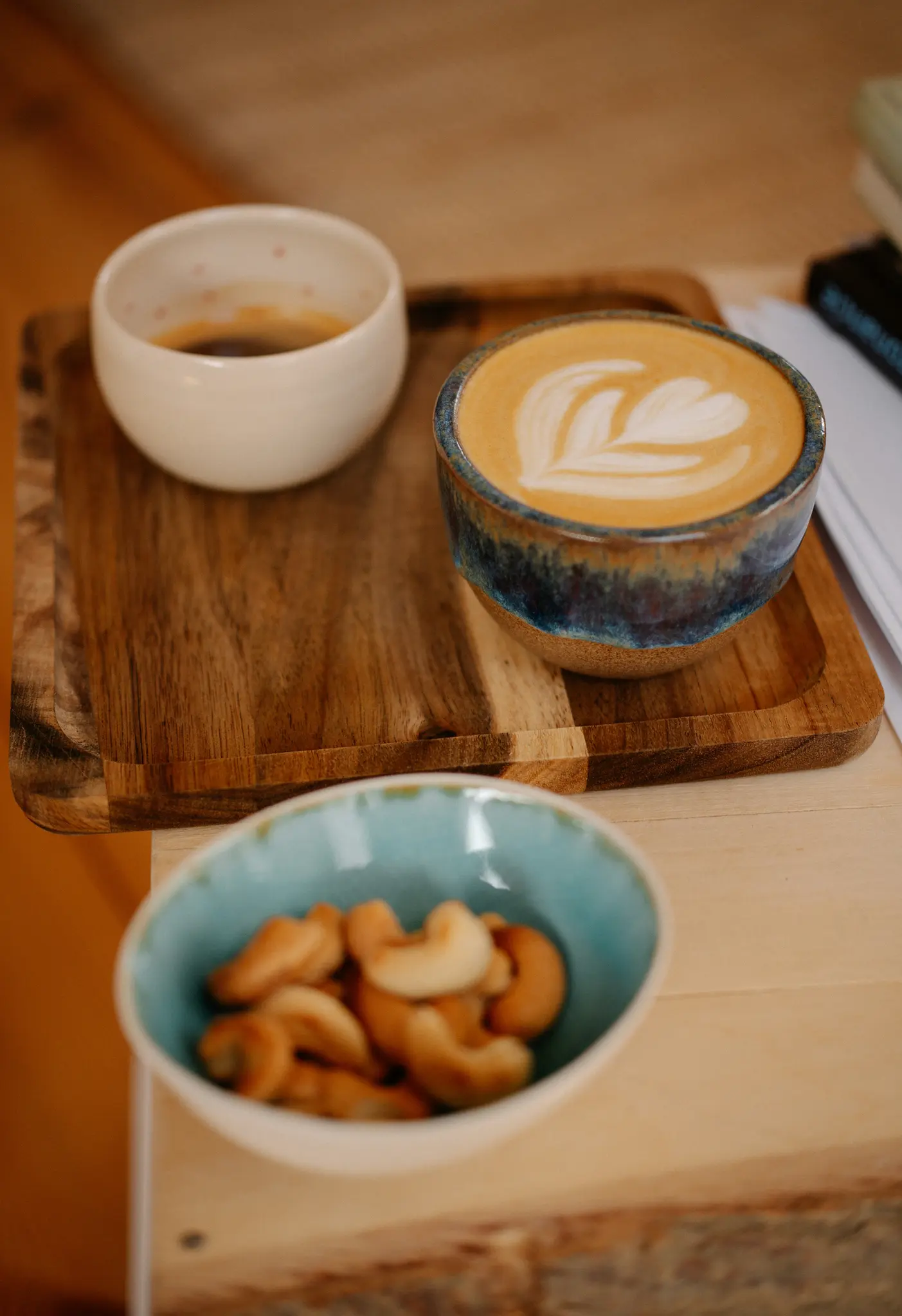 Two cups of coffee and a bowl of cashews are arranged on a wooden tray. One cup is white with black coffee, the other is…