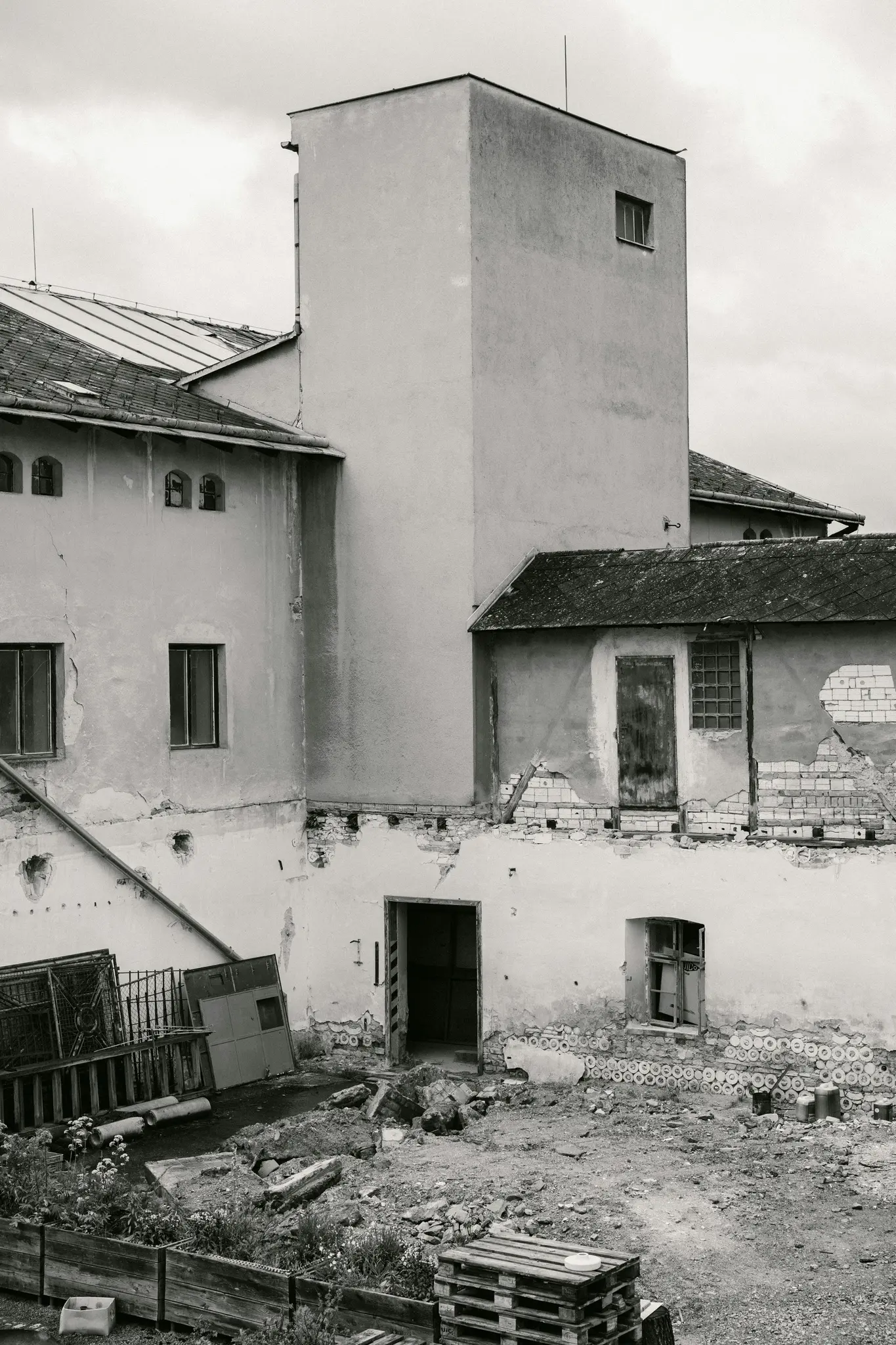 Černobílá fotografie zobrazuje vícepodlažní komplex budov s vysokou, prostou věží a rozpadajícími se zdmi, obklopený...