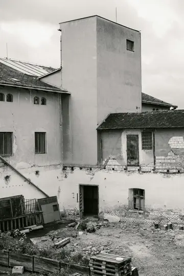 Černobílá fotografie zobrazuje vícepodlažní komplex budov s vysokou, prostou věží a rozpadajícími se zdmi, obklopený...