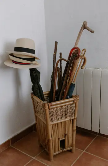A wicker stand holds two straw hats, a black umbrella, a blue umbrella, and several walking sticks and canes.