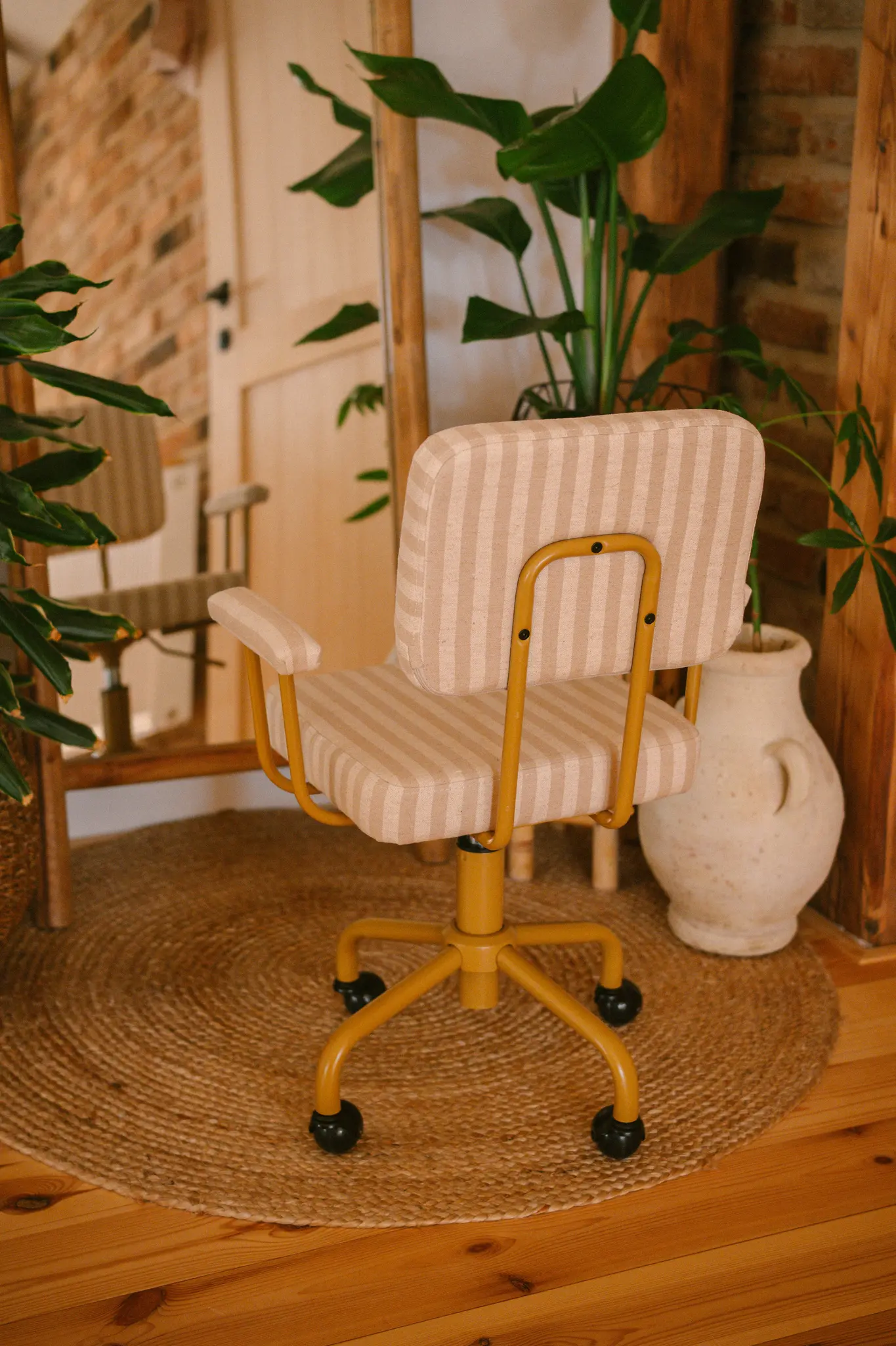 Een mosterdgele bureaustoel met beige en wit gestreepte bekleding staat op een rond jute vloerkleed. Potplanten en een