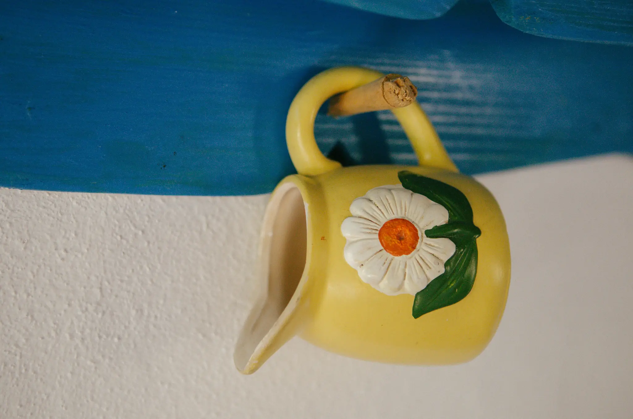 Een gele keramische kan met een wit en oranje madeliefjesontwerp hangt aan een blauw houten oppervlak tegen een witte muur.