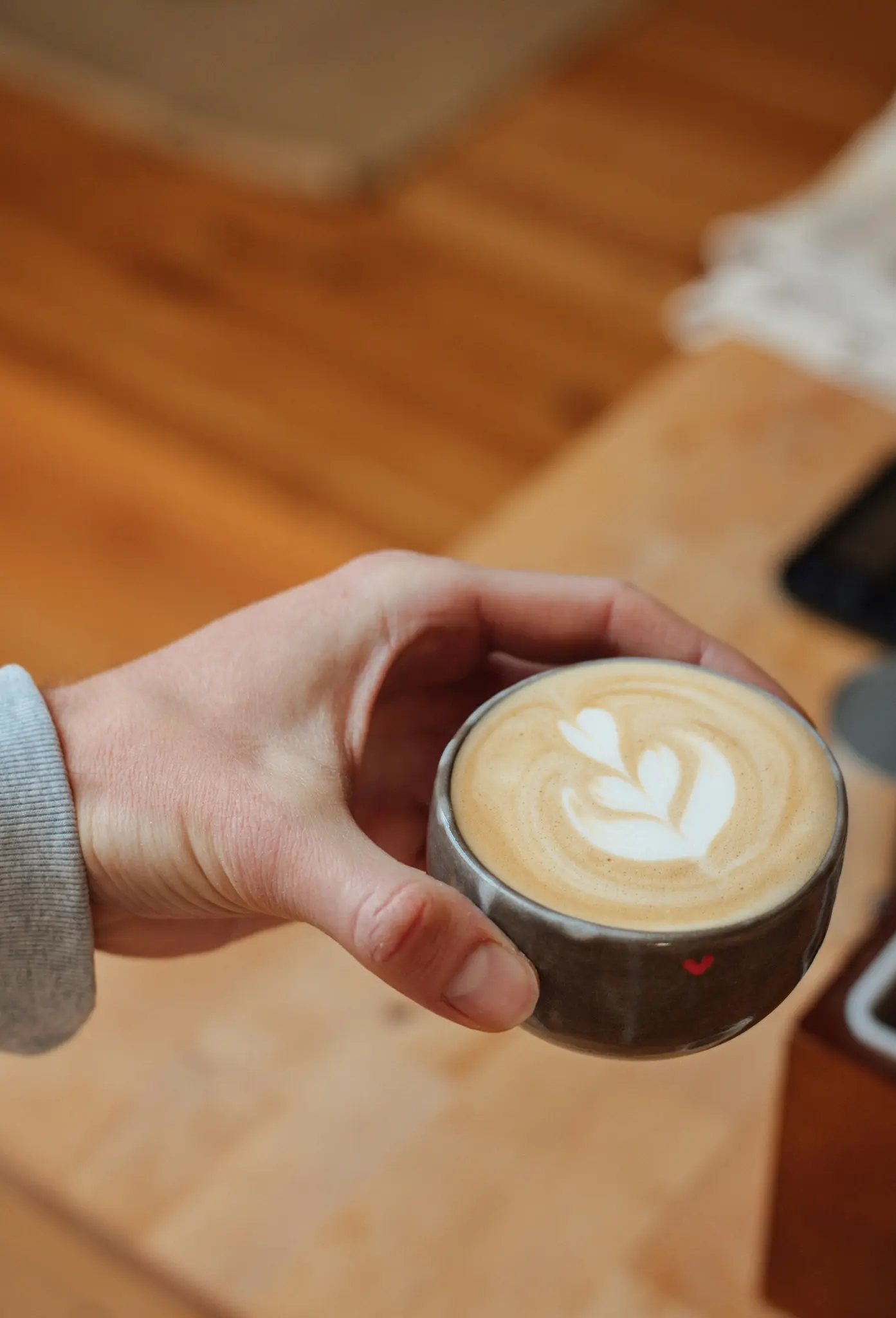 En hand i en grå ärm håller en mörkgrå keramisk mugg fylld med latte. Latten har vit skumkonst i hjärtform.