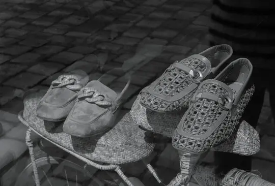 Due paia di scarpe, una in camoscio e una intrecciata, sono esposte su supporti intrecciati in una foto in bianco e nero.