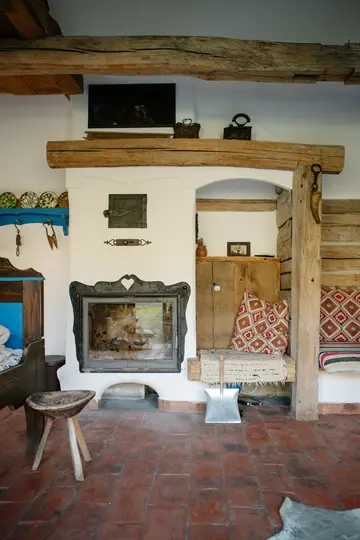 Een rustieke kamer heeft een witte open haard met een zwarte metalen deur, omgeven door zware houten balken. Terracotta