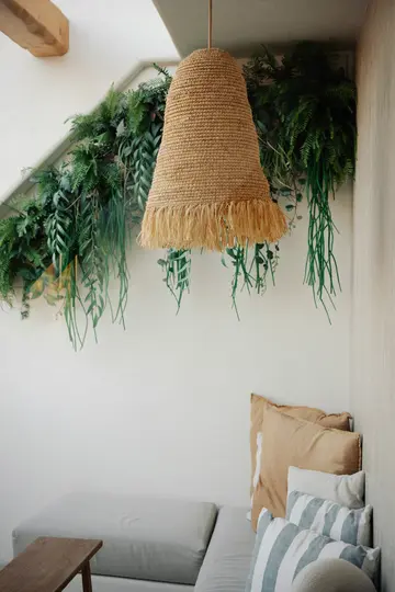 Een geweven strooien lampenkap met franjes hangt aan het plafond. Daaronder een witte bank met kussens in beige en blauw.