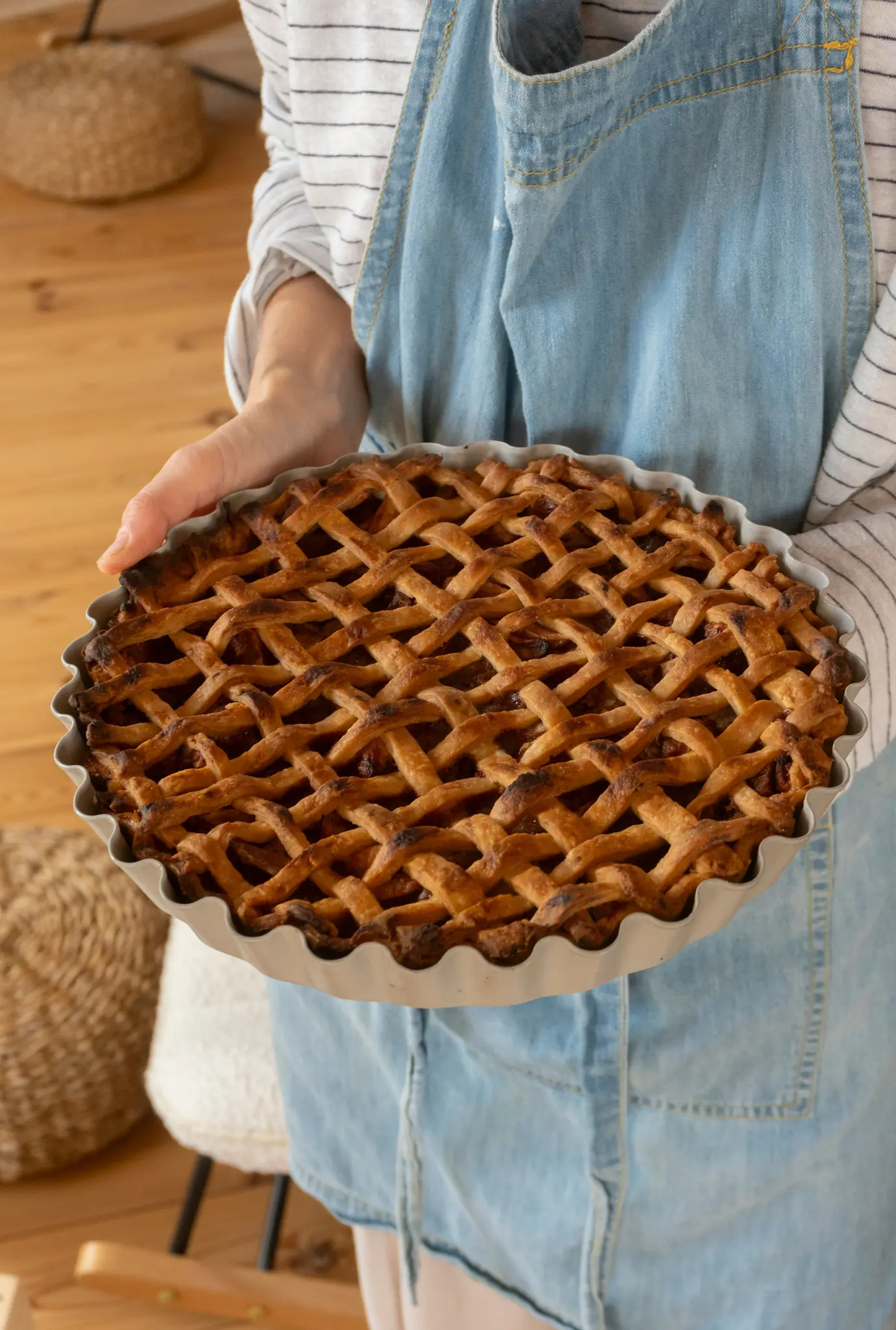 Une personne portant un tablier en jean et une chemise rayée tient une tarte dorée avec un dessus en treillis dans un moule