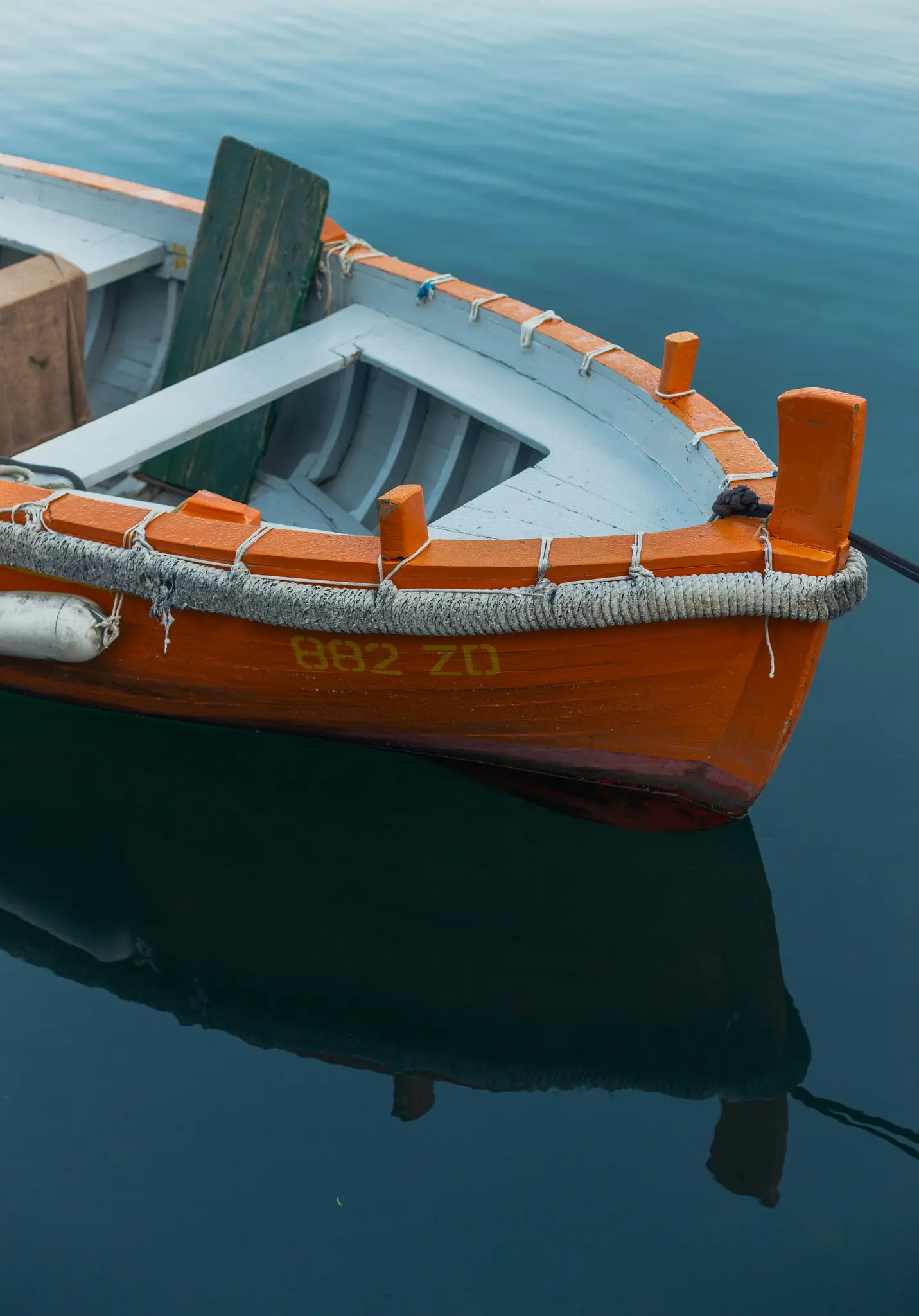 Een houten vissersboot met een oranje romp en een lichtblauwe binnenkant drijft op stil, donkerblauw water. De boot heeft een