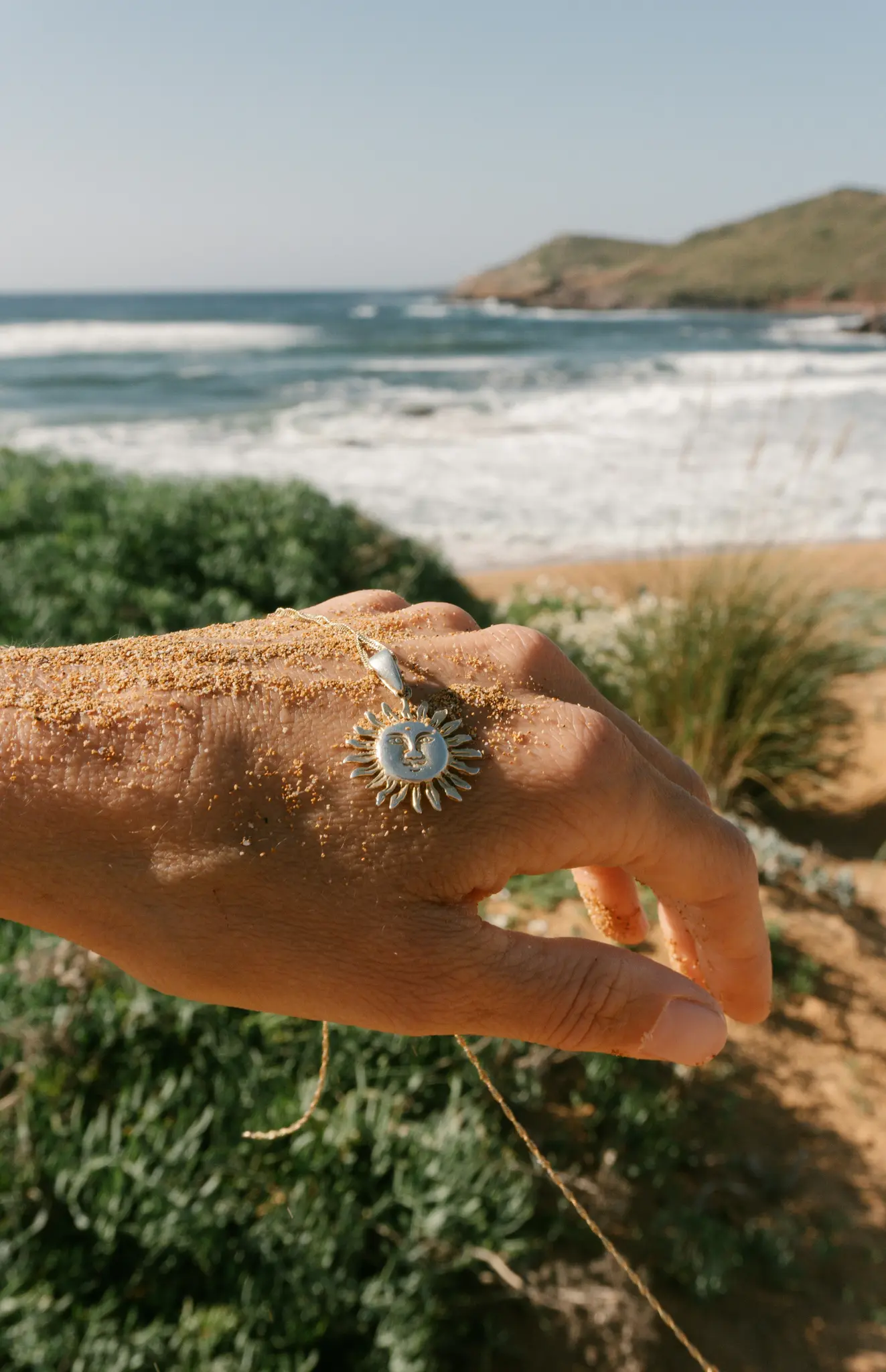 Nahaufnahme einer sandbedeckten Hand, die einen silbernen, sonnenförmigen Anhänger mit einem Gesicht hält, vor einem