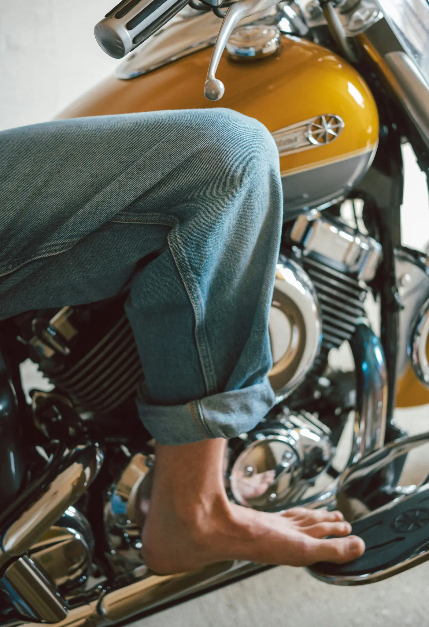 Een been in een spijkerbroek en een blote voet rustend op de voetsteun van een gele motorfiets met chromen details.