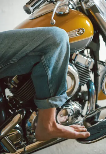 Noha v modrých džínách a bosá noha spočívající na stupačce žlutého motocyklu s chromovými detaily.