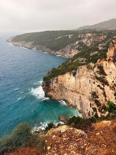 Vista dall'alto di una costa frastagliata con scogliere ripide e chiare coperte da vegetazione verde. Il mare turchese…