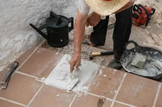 Una persona con cappello di paglia e pantaloni neri applica stucco bianco su piastrelle quadrate marroni con una cazzuola.