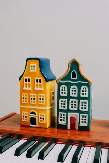 Two colorful ceramic house-shaped candle holders, one yellow and one teal, sit on a wooden surface above a piano keyboard.