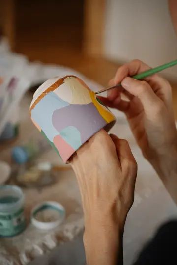 Nærbillede af hænder, der maler et keramikkrus. Kruset er dekoreret med abstrakte former i pastelfarver som pink, blå og...