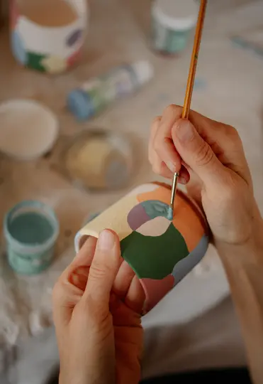 Nærbillede af hænder, der maler en keramikkop med en pensel og påfører lyseblå maling på et farverigt abstrakt design.