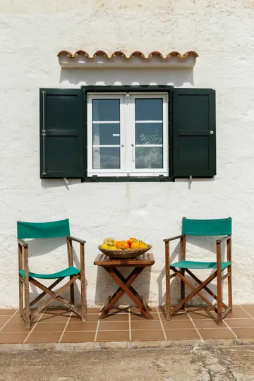 Un muro in stucco bianco presenta una finestra con persiane verde scuro. Sotto, due sedie di legno con tessuto verde