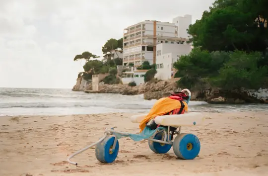 Una carrozzina da spiaggia blu con ruote grandi si trova su una spiaggia sabbiosa, drappeggiata con un asciugamano arancione