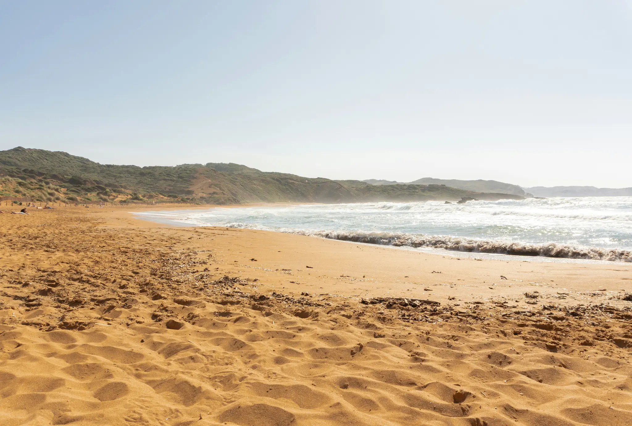 Un'ampia inquadratura di una spiaggia sabbiosa dorata con onde spumeggianti che si infrangono sulla riva. Colline verdi