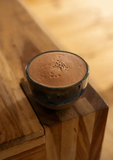 A close-up shot of a dark blue and brown ceramic bowl filled with a frothy, light brown beverage, resting on a wooden…