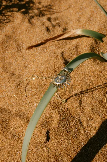 Un ciondolo d'argento a forma di sole con un volto stilizzato poggia su una foglia di un verde vibrante, posizionata su