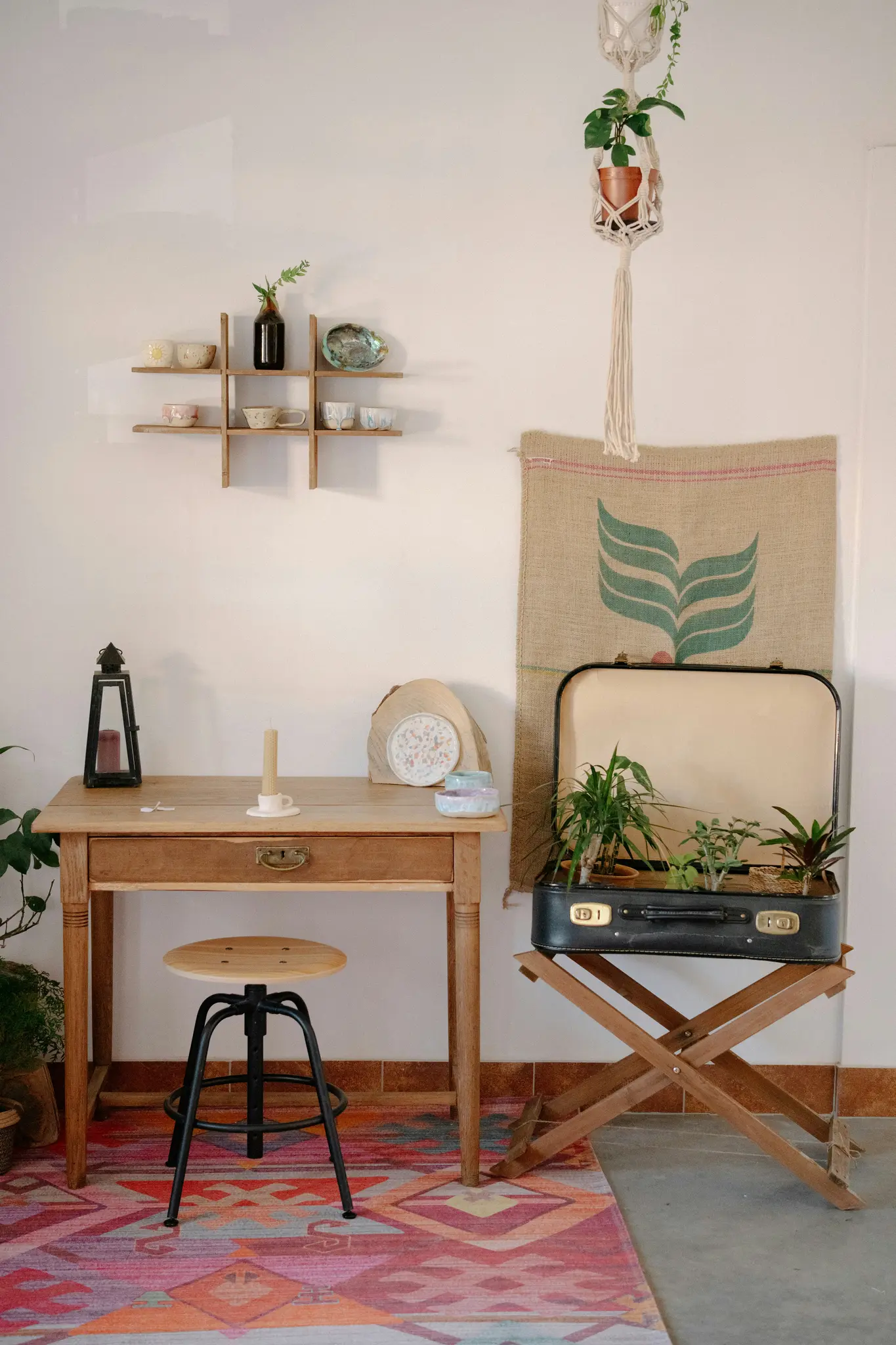 Een gezellige binnenruimte met een houten bureau, een zwarte kruk en een vintage koffer vol planten. Een kleurrijk tapijt