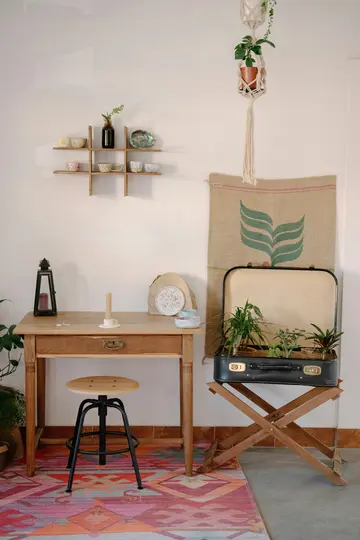 Útulná vnitřní scéna s dřevěným stolem, černou stoličkou a vintage kufrem plným rostlin. Na podlaze je barevný koberec.