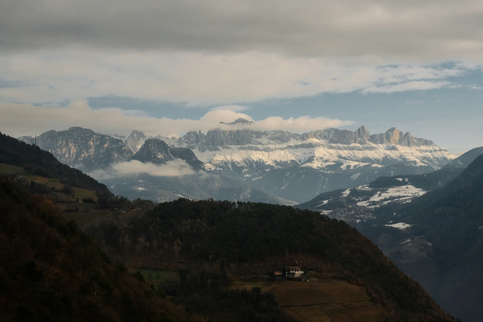 Een brede landschapsfoto met sneeuwbedekte bergen onder een bewolkte hemel, met donkere beboste heuvels en een vallei in de…