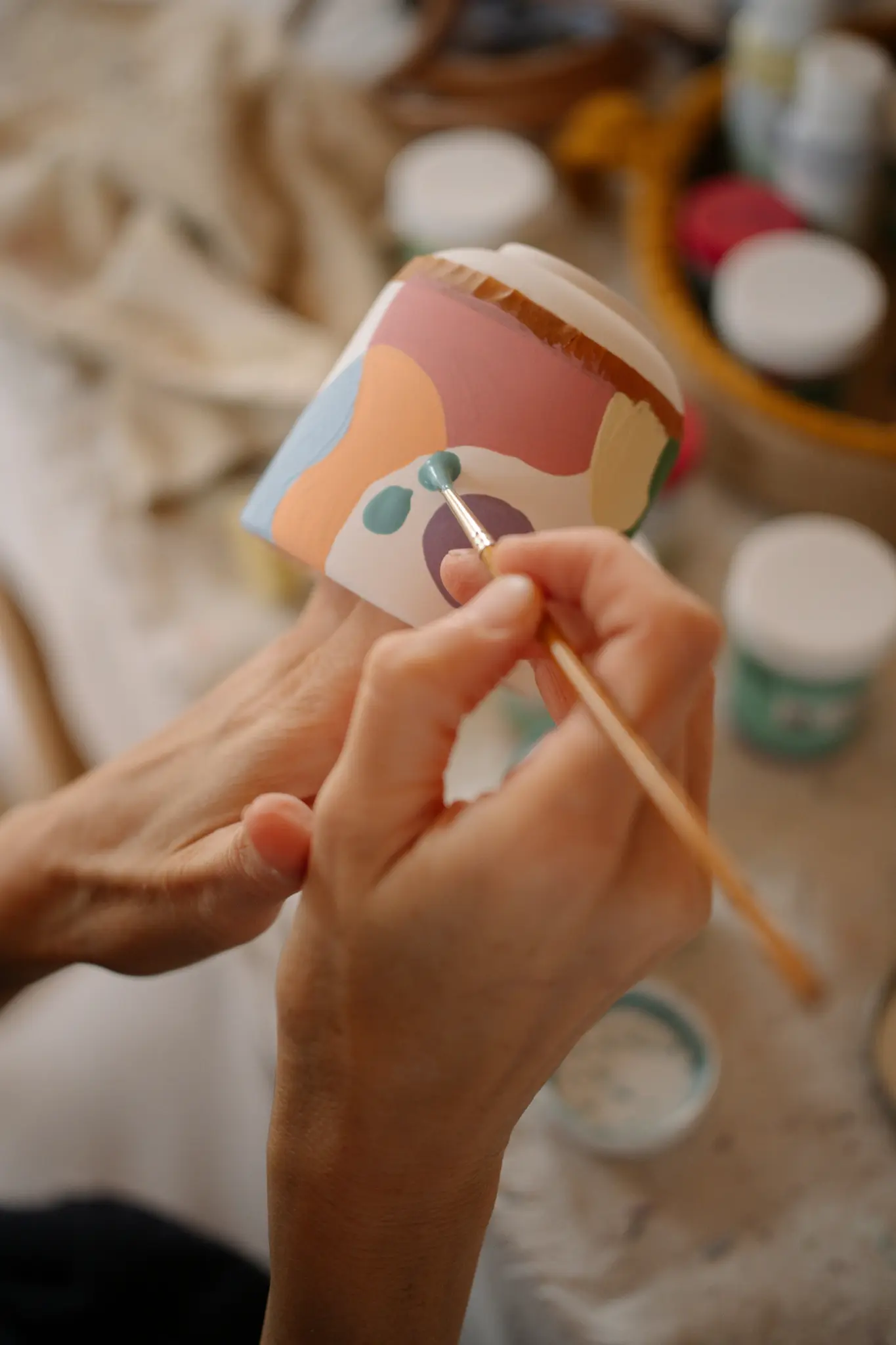 Gros plan sur des mains peignant un pot en céramique avec des formes abstraites dans des couleurs pastel. Un petit pinceau
