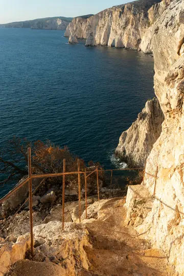 Un chemin escarpé et sinueux en roches claires et en terre descend vers la mer d'un bleu profond. Une rambarde…