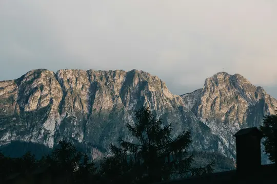 Una maestosa catena montuosa sotto un cielo morbido e nuvoloso, con la luce del sole che illumina le cime rocciose. Alberi