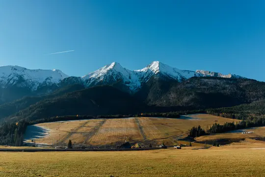 Široká krajinářská fotografie zobrazující zasněžené hory v pozadí, tmavě zelené zalesněné kopce a zlato-hnědá pole.