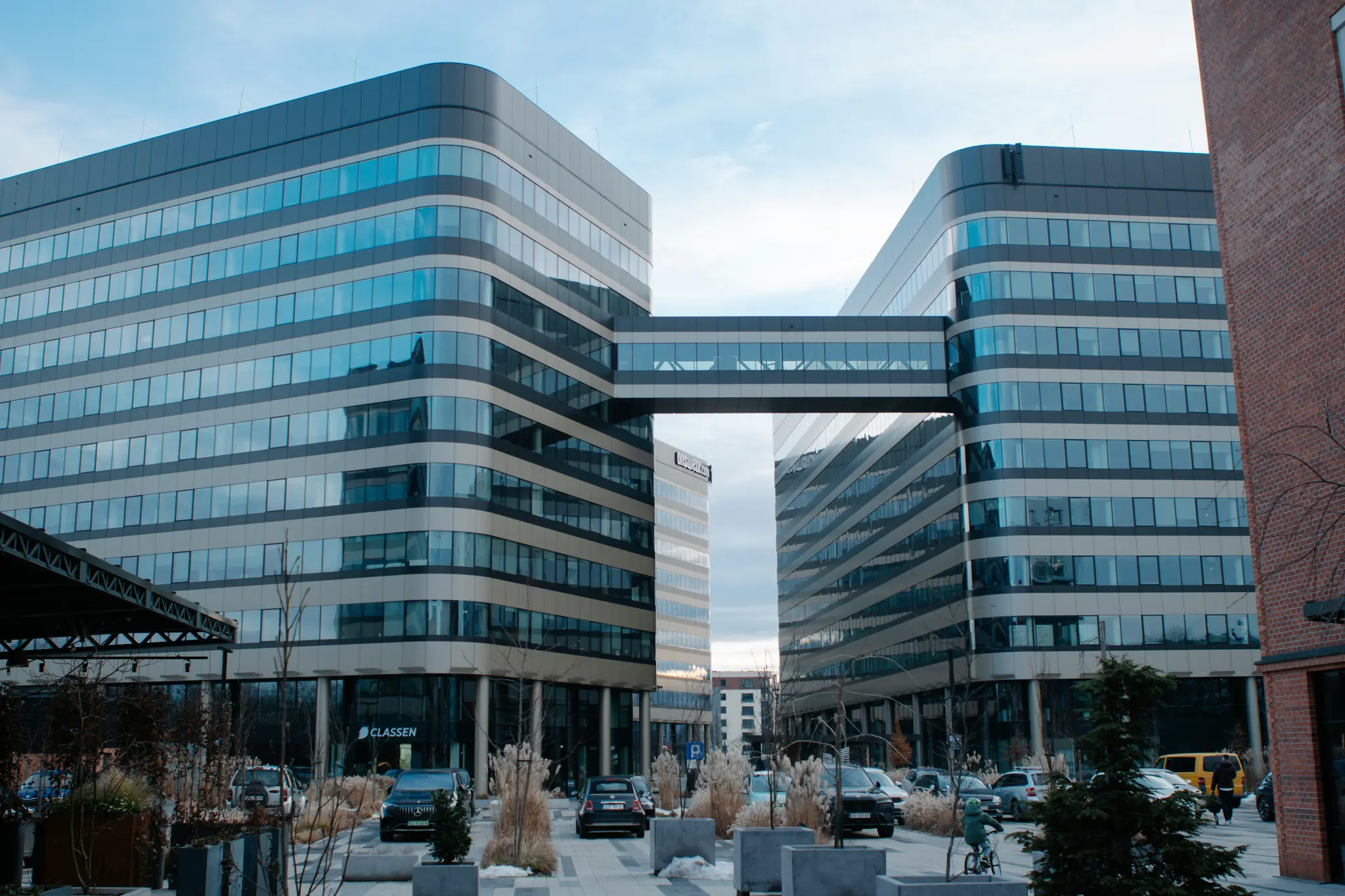 En vy över två moderna kontorsbyggnader med böjda fasader och en skybridge som förbinder dem, sedd från en stenlagd