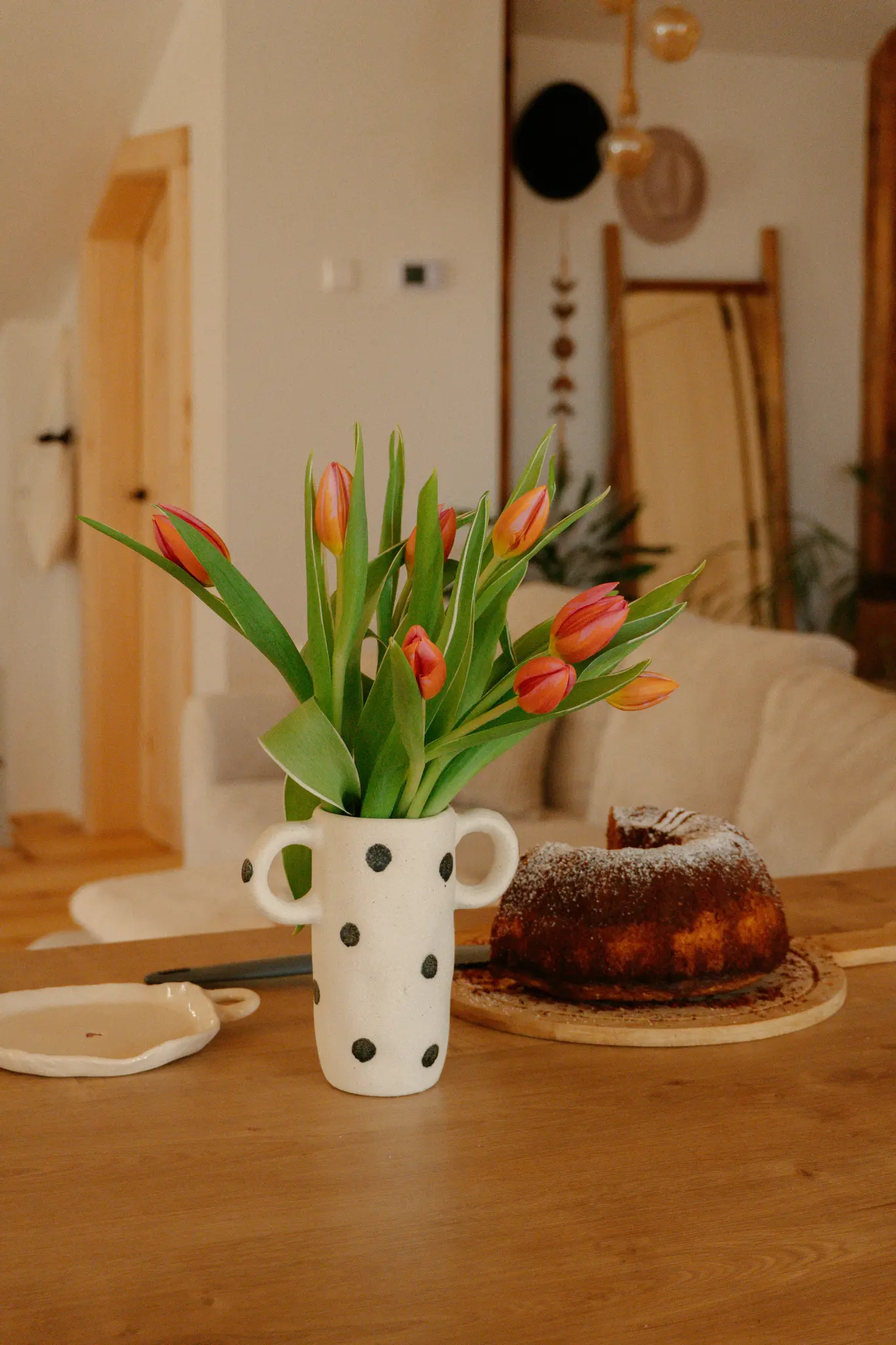 Een witte vaas met zwarte stippen bevat een boeket oranje en rode tulpen naast een bundt cake bestrooid met poedersuiker…