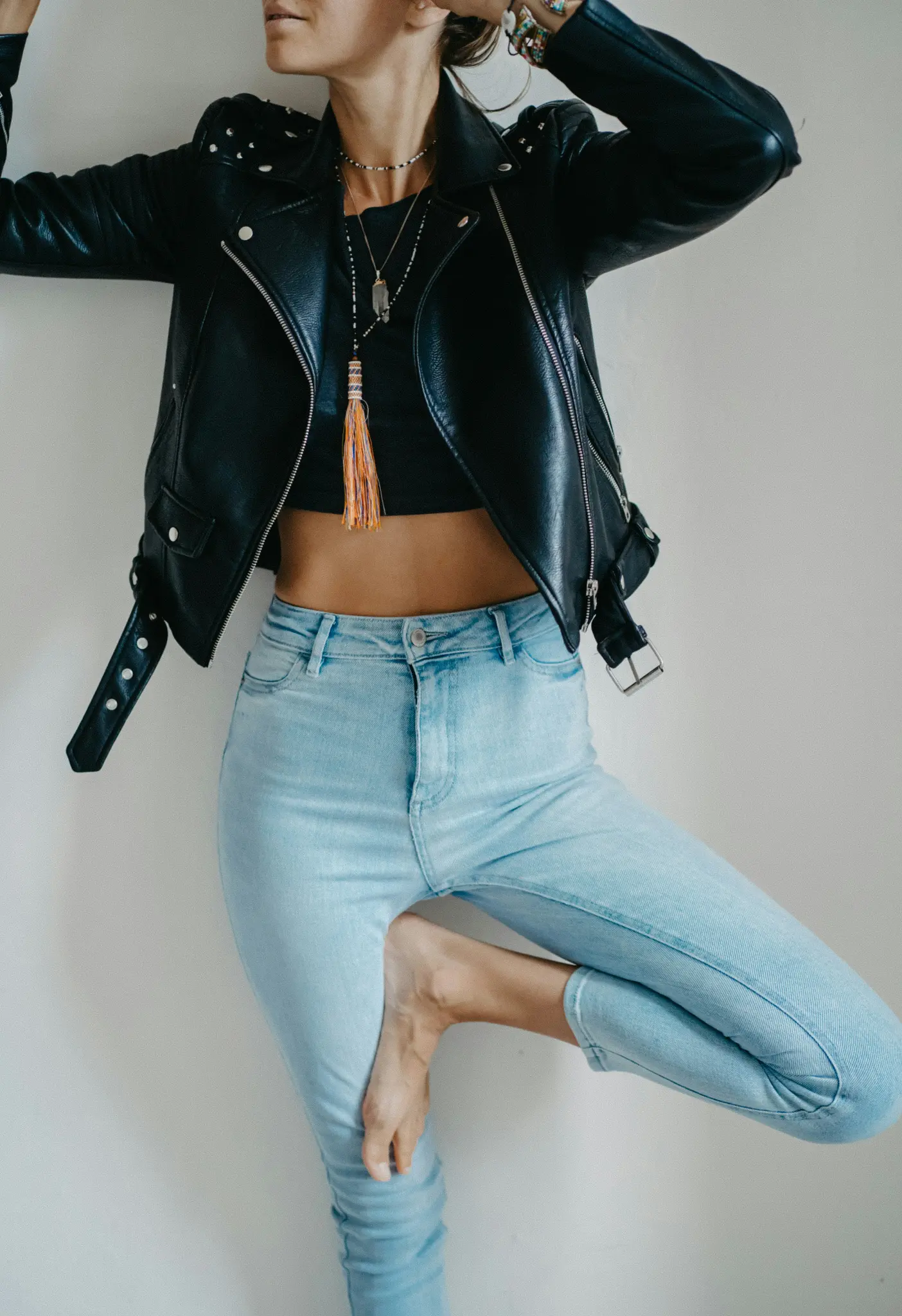 Eine Person in einer schwarzen Lederjacke, einem schwarzen Crop-Top und hellblauen Jeans steht in der Baum-Yoga-Pose vor