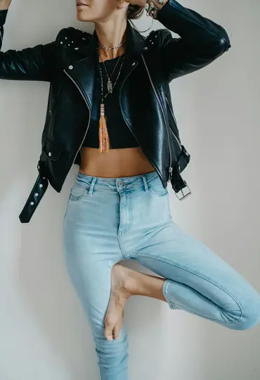 En person i en sort læderjakke, sort crop top og lyseblå jeans står i en yogatræstilling op ad en hvid væg.