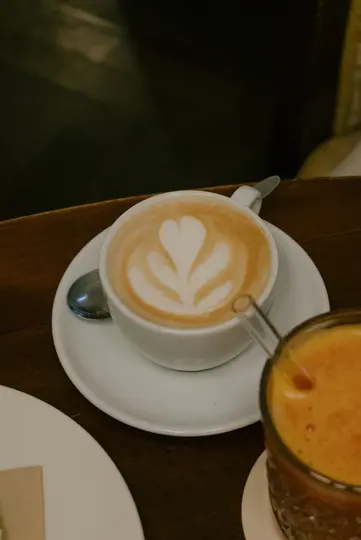 A white cup of latte with a heart-shaped latte art and a spoon on a saucer, next to a glass of orange juice with a straw…