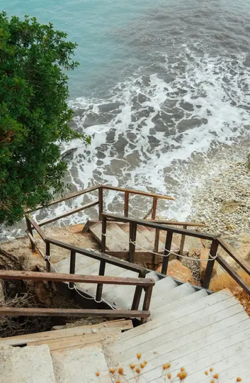 Vista dall'alto di una scala tortuosa con ringhiere in legno che scende verso una spiaggia rocciosa con onde spumeggianti.