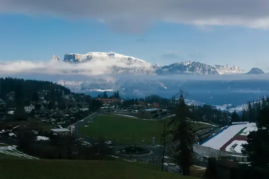 A wide landscape photo shows a village nestled in a valley with snow-capped mountains partially obscured by clouds in the…