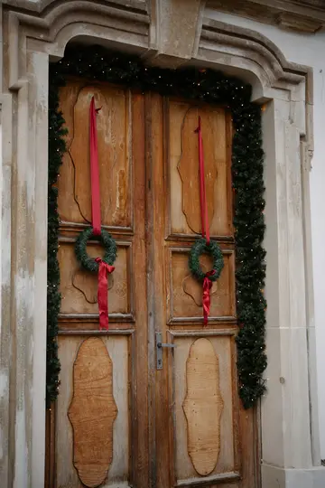 A rustic wooden double door with a stone frame is adorned with green garlands and small lights. Two green wreaths with red…