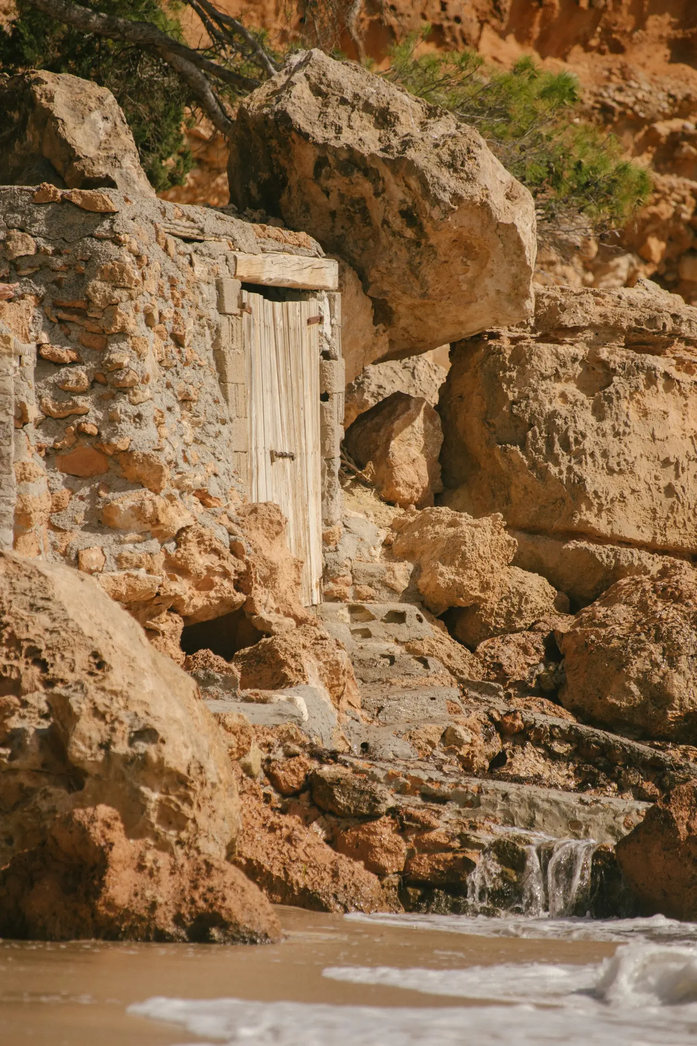 Eine rustikale Holztür ist in eine raue, orangebraune Felswand eingelassen, mit kleinen Stufen, die zu ihr führen. Wasser