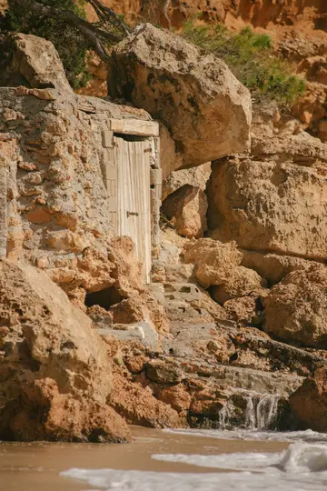 Una porta rustica in legno è incastonata in una scogliera rocciosa aspra e marrone-arancio, con piccoli gradini che conducono